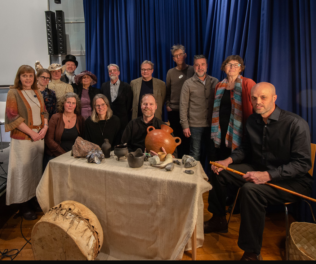 Gruppfoto med sju kvinnor och sju män samlade bakom ett bord med stenåldersinspirerade instrument.