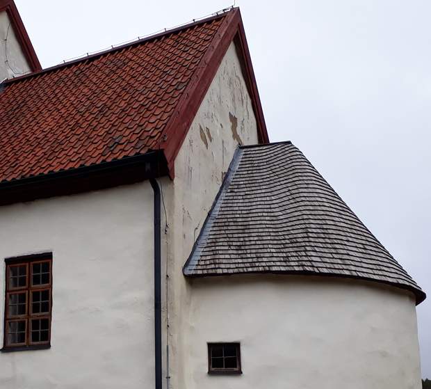 Färgfoto av taket på absiden klätt med gråbruna träspånor. Korets tak, intill, är klätt med rött tegel.