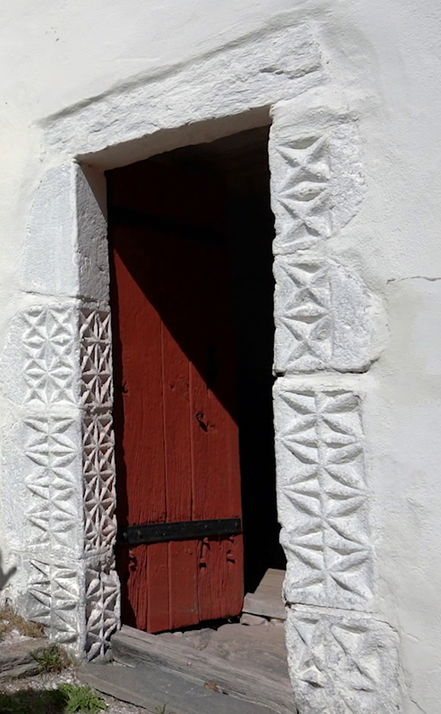Färgfoto på port med omgivande vägg i vit kalksten. Portens omfattning är dekorerad med ett inskuret geometriskt mönster.