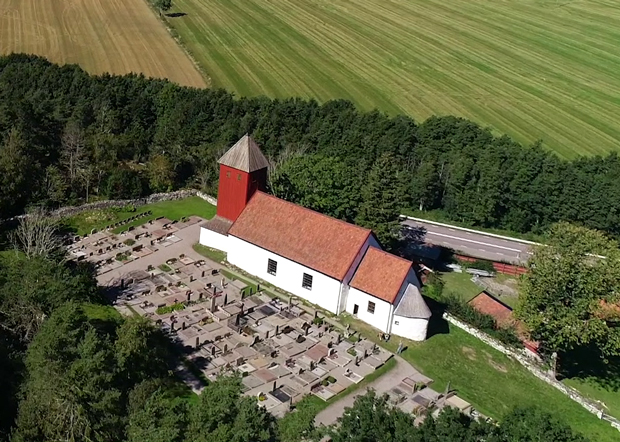 Flygfoto i färg över kyrkan och kyrkogården omgivna av träddungar och åker. 