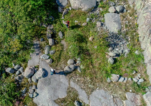 Foto. Ett stenigt sommarlandskap sett ur fågelperspektiv. Berg i dagen mellan gräsmark och lövträd. Bumlingar av granit ligger i en oregelbunden formation. 