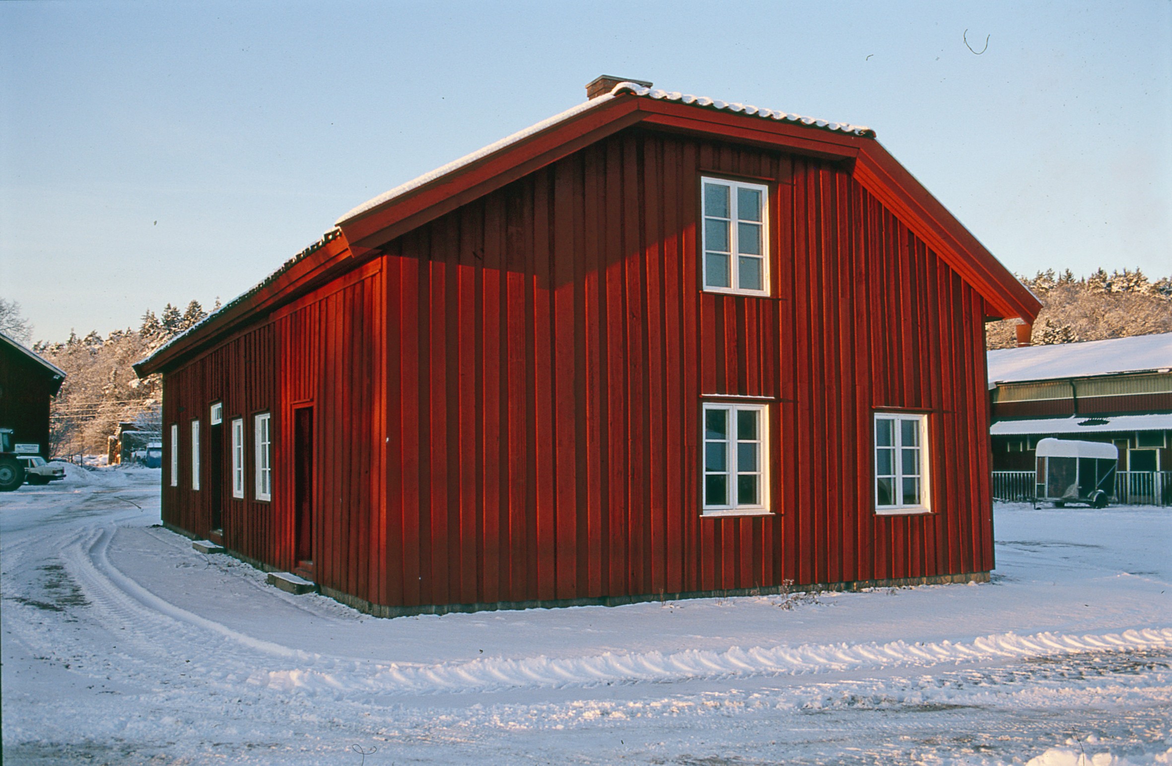 Färgfoto visar röd byggnad med vita fönster på en snötäckt gårdspan.