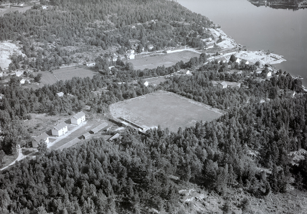 Svartvit flygbild med fotbollsplan och några hus, omgivet av skog. Fjorden syns längst upp till höger.
