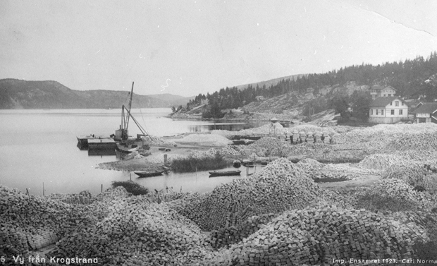 Svartvitt foto visar stora högar med gatsten. Fjorden, några båtar och ett par hus i bakgrunden.