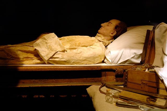 Installation. En skulptur föreställande Karl den tolfte som lik. Han vilar på en brits av trä och är klädd i vit skjorta.