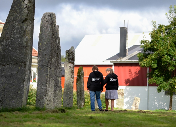 Färgfoto med fyra stycken 2-4 meter höga resta stenar på rad, två personer och en ladugård i bakgrunden.