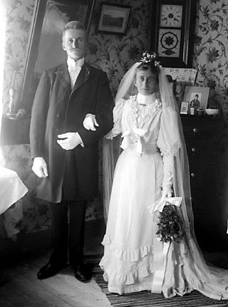 Svartvitt foto av brudpar i hemmet. Bruden har vit klänning med hög hals och fårbogsärmar samt slöja och krona.