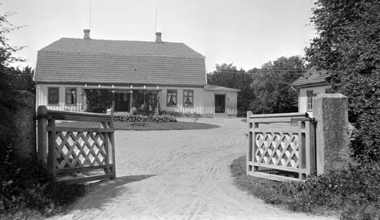 Svartvitt foto visar huvudbyggnad med brutet tak, gårdspan och en öppen grind i förgrunden.