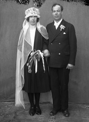 Svartvitt foto av brudpar. Brudgummen i mörk kostym, bruden har svart knälång dräkt med lång vit slöja som täcker huvudet och är lång neddragen i pannan.