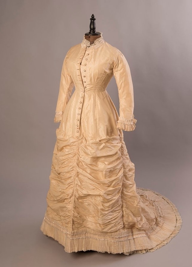 Färgfoto av gulvit klänning med knäppning fram och draperad kjol med släp.