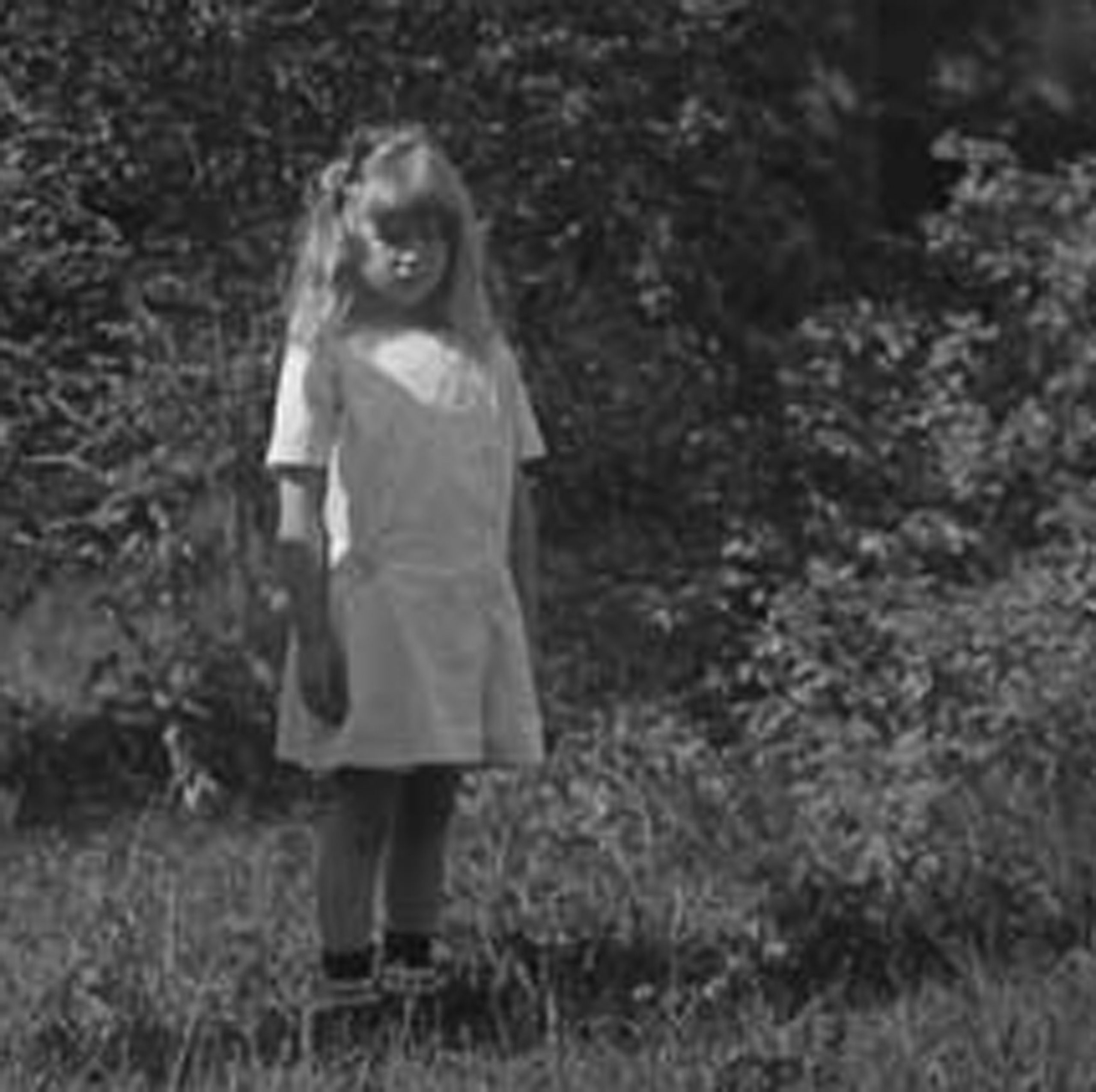 Svartvitt foto. En flicka cirka 5 år gammal med utsläppt hår och bara ben står i en trädgård.