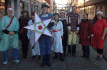 Foto. Nio medelålders personer utklädda i julinspirerad klädsel. I mitten står en man med en julstjärna i vitt. De står utomhus på Kungsgatan i Uddevalla