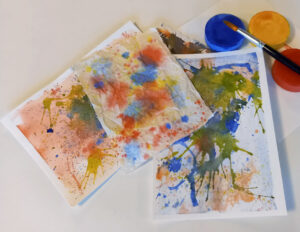 Foto. Papper med färgstänk på mot vit bakgrund. Till vänster röd, blå, gul rund kaka med vattenfärg och en pensel.