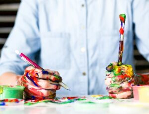foto. En person sitter med färgiga händer. En penna i ena handen och en pensel i andra.