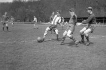 Svartvitt foto. En fotbollsmatch. En ung man i randigt försöker dribbla sig förbi två spelare i helfärgade tröjor, En av försvararna har en keps.